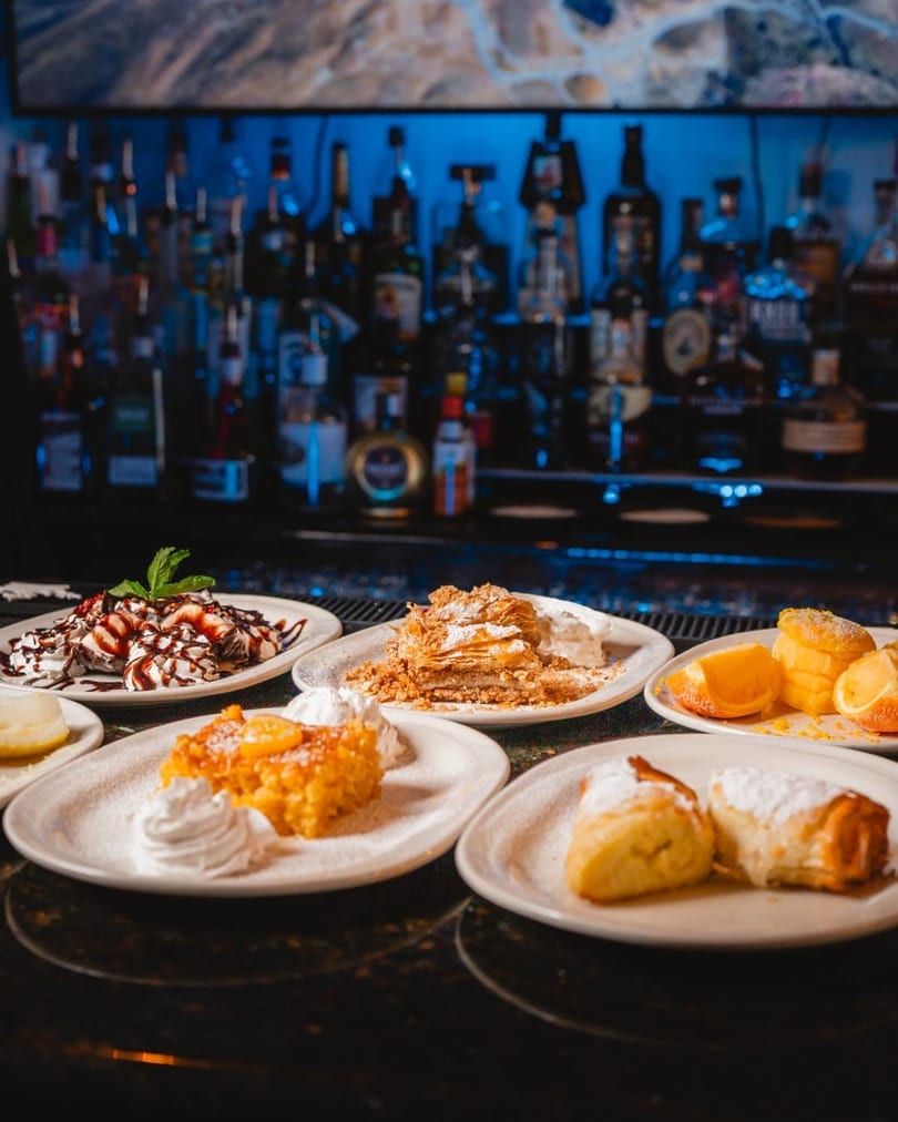 Assortment of desserts including baklava, cakes, and pastries on a bar with liquor bottles.