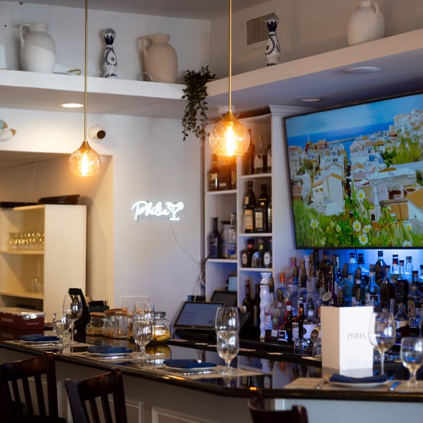 A well-stocked bar with a neon "Philia" sign, a large TV, and set tables with blue napkins.