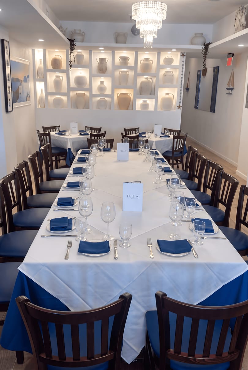 A long dining table set with white tablecloths, blue napkins, and wine glasses in a bright room.