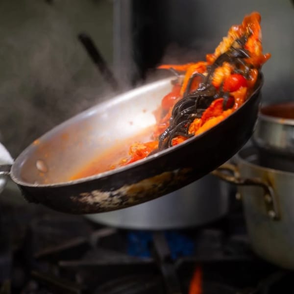 Chef tossing black pasta with red sauce and cherry tomatoes in a pan over a gas flame.