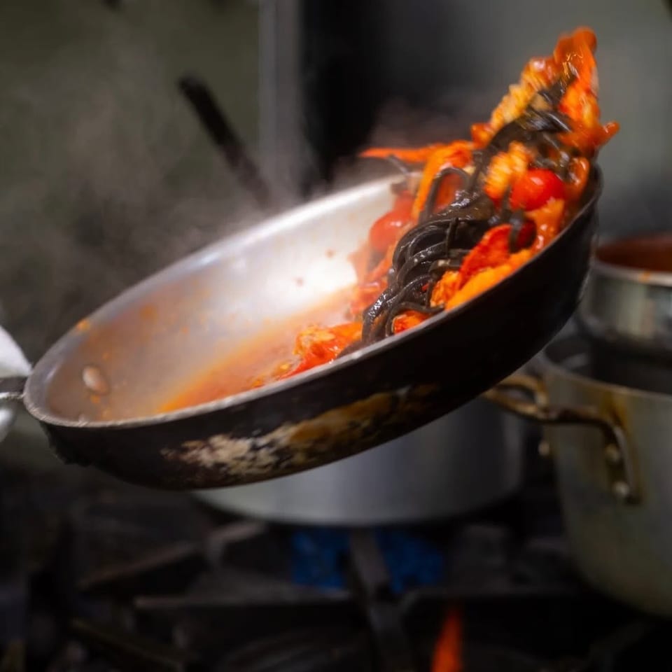 Chef tossing black pasta with red sauce and cherry tomatoes in a pan over a gas flame.