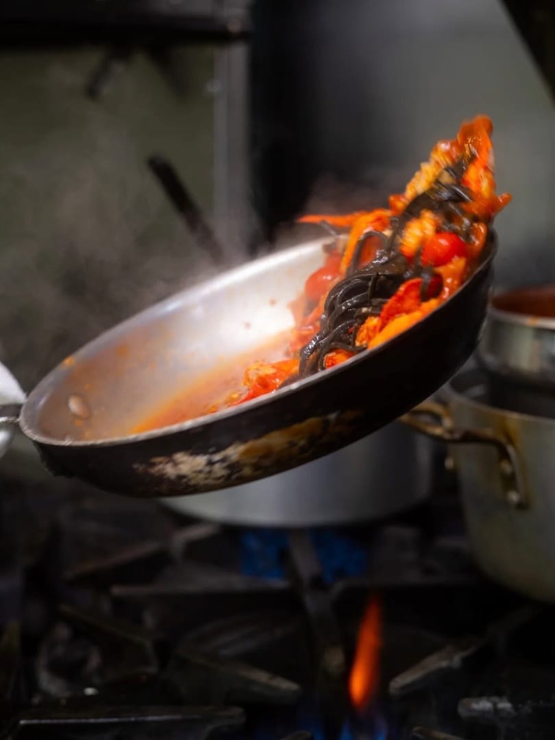 Chef tossing black pasta with red sauce and cherry tomatoes in a pan over a gas flame.