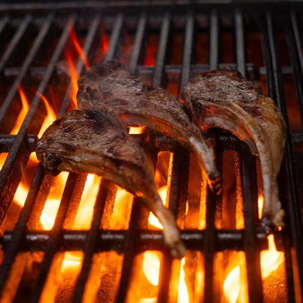 Three lamb chops sizzle on a fiery grill, flames licking up through the grates.