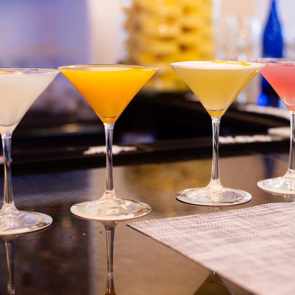 Four colorful martini cocktails, including white, orange, yellow, and pink, lined up on a bar.