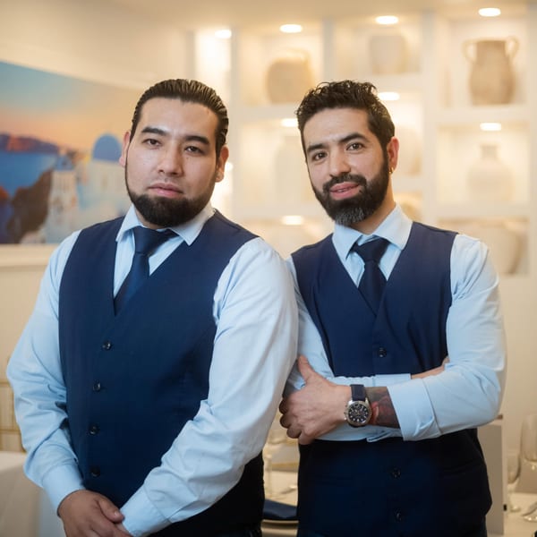 Two men in blue vests and ties stand in a restaurant with white decor and art.