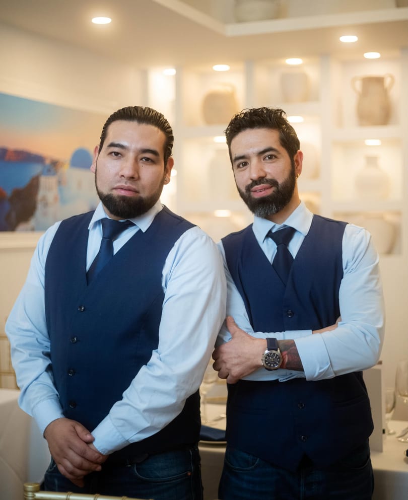 Two men in blue vests and ties stand in a restaurant with white decor and art.