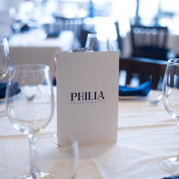 A white menu for Philia Restaurant stands on a table set with wine glasses and blue napkins.