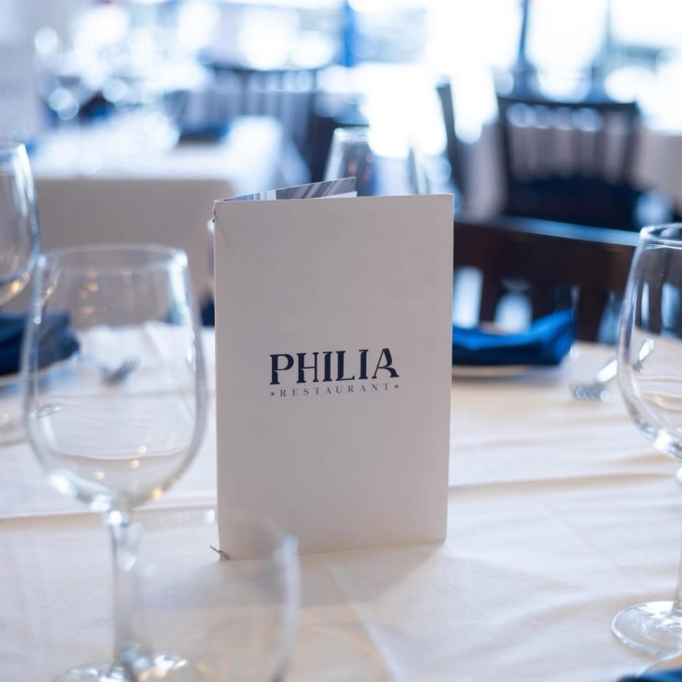 A white menu for Philia Restaurant stands on a table set with wine glasses and blue napkins.