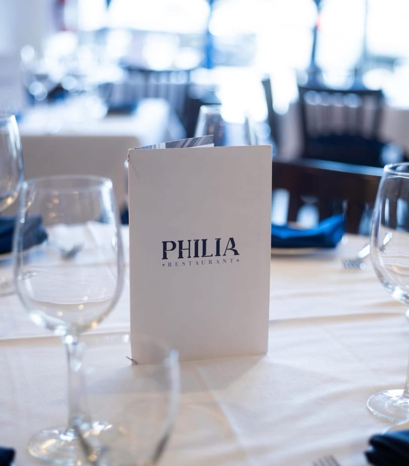 A white menu for Philia Restaurant stands on a table set with wine glasses and blue napkins.