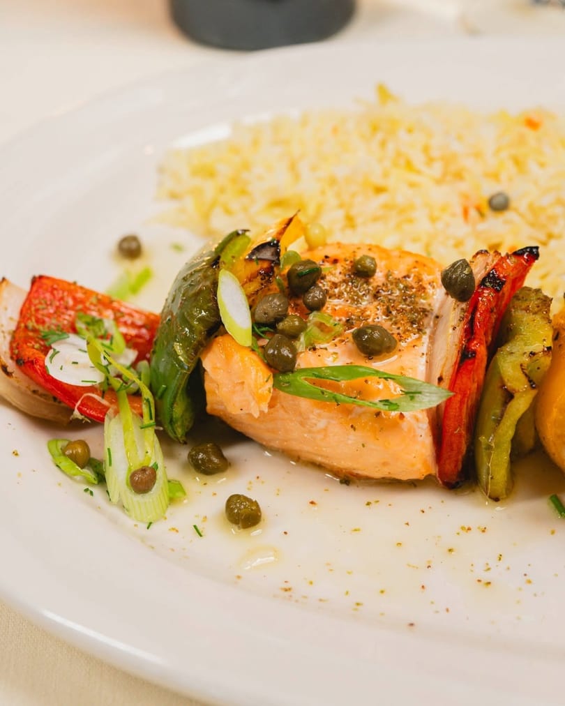 Grilled salmon skewer with bell peppers, onions, capers, and rice on a white plate.