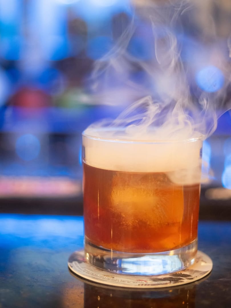 A smoky cocktail with a large ice cube sits on a coaster against a blurred blue background.