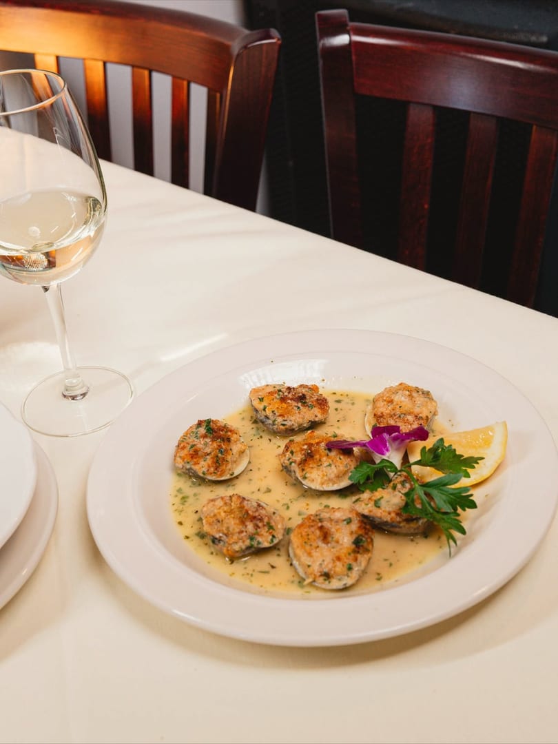 Baked clams in a creamy sauce with lemon and parsley, next to a glass of white wine.