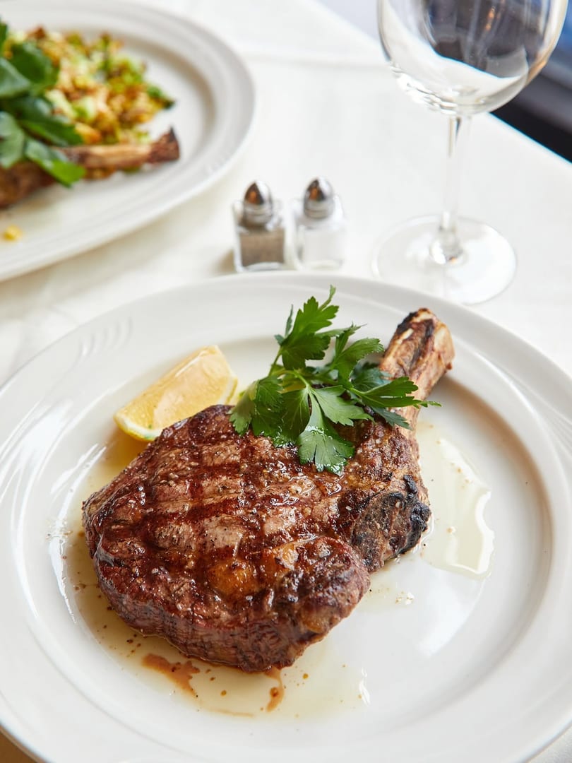 Grilled steak with a bone, garnished with parsley and a lemon wedge on a white plate.