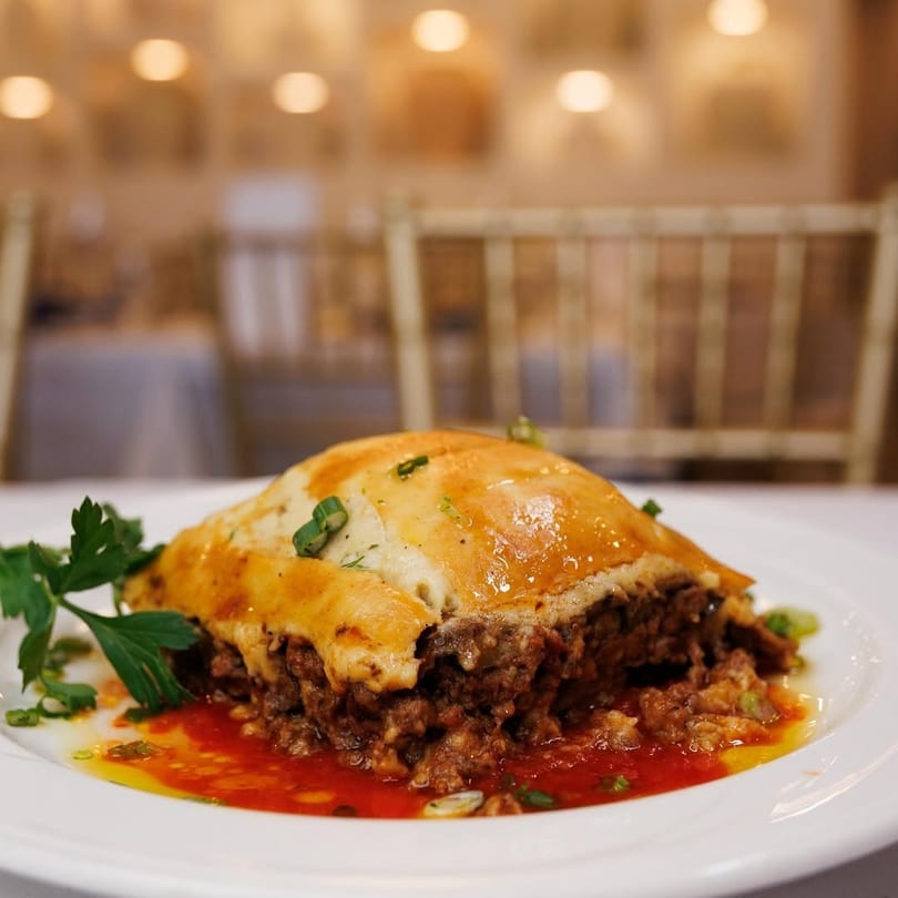 A serving of moussaka with a golden-brown top, rich meat filling, and red sauce on a white plate.