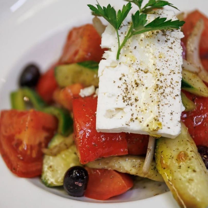 Close-up of a vibrant Greek salad with a large feta cheese block, olives, and fresh vegetables.