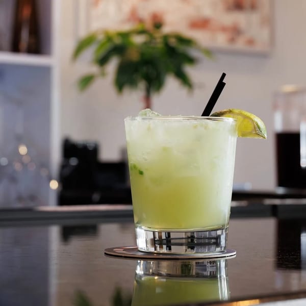 Lime green cocktail with ice, a lime wedge, and black straw on a dark reflective bar.