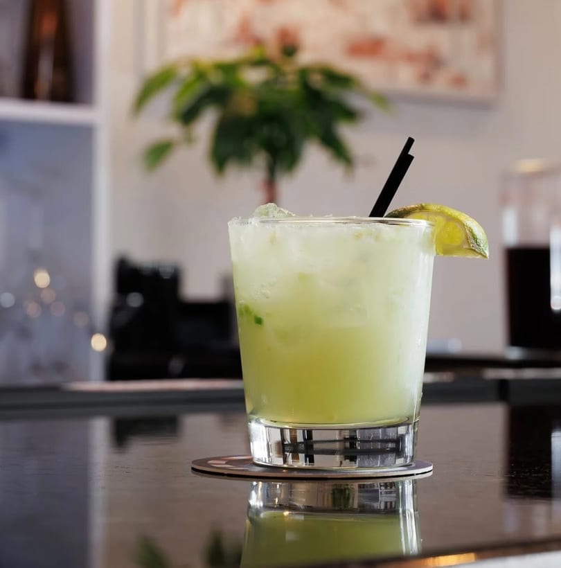 Lime green cocktail with ice, a lime wedge, and black straw on a dark reflective bar.