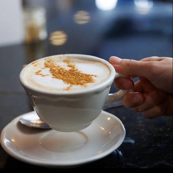 A hand holding a white cup of cappuccino with cinnamon sprinkled on top, on a dark table.