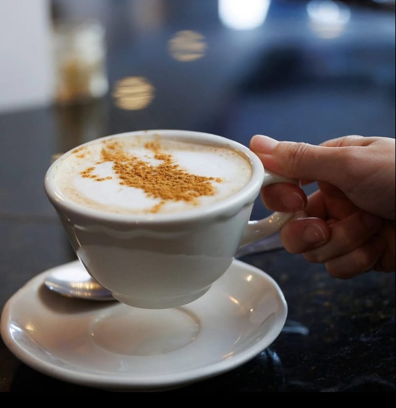 A hand holding a white cup of cappuccino with cinnamon sprinkled on top, on a dark table.