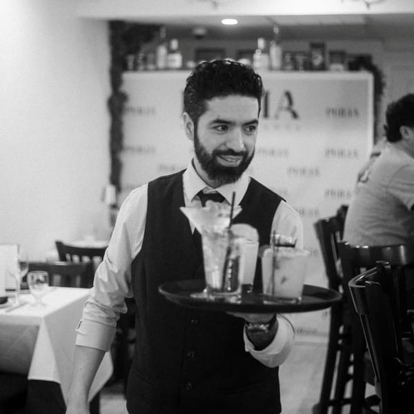Smiling waiter in a vest carrying a tray of drinks through a restaurant dining room.