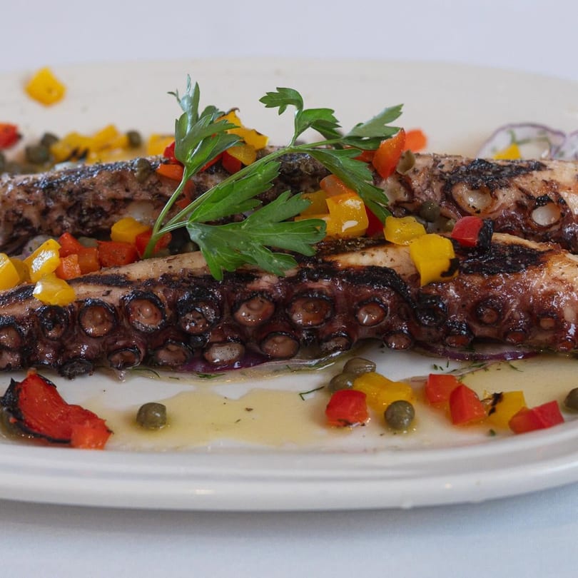 Grilled octopus with chopped red and yellow bell peppers, capers, and parsley on a white plate.