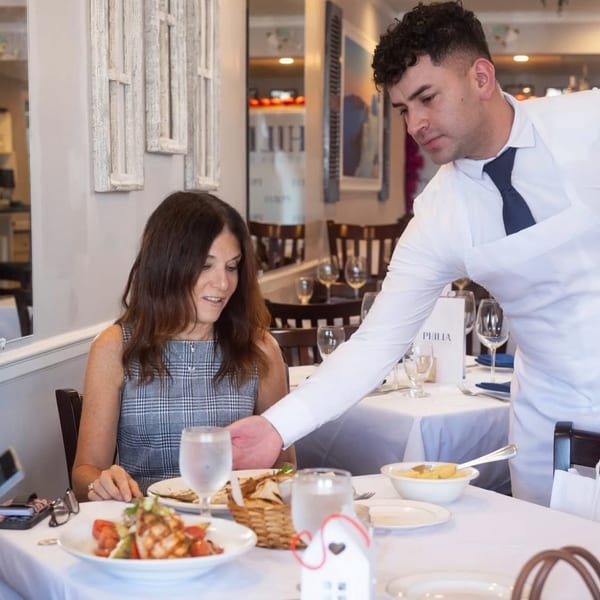A server in a white shirt and blue tie serves a woman dining at a restaurant table.
