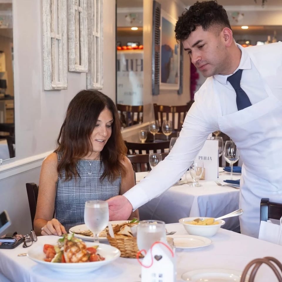 A server in a white shirt and blue tie serves a woman dining at a restaurant table.