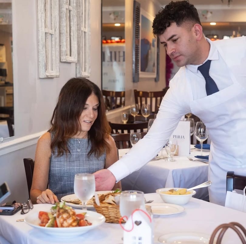 A server in a white shirt and blue tie serves a woman dining at a restaurant table.