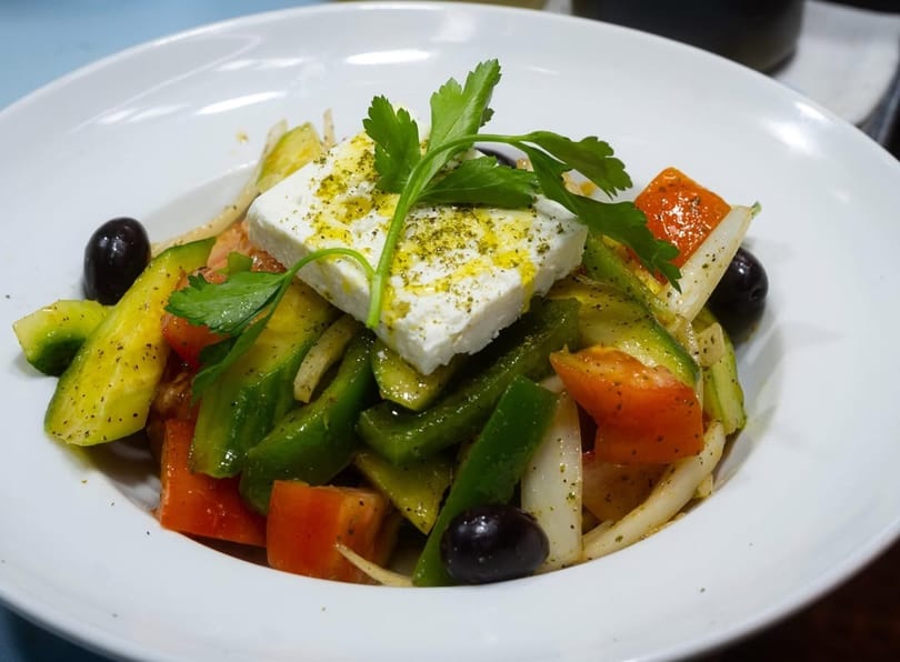 A vibrant Greek salad with feta, olives, tomatoes, cucumbers, and bell peppers in a white bowl.