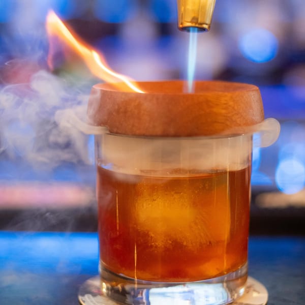 A bartender uses a torch to smoke a cocktail in a glass with a large ice cube.