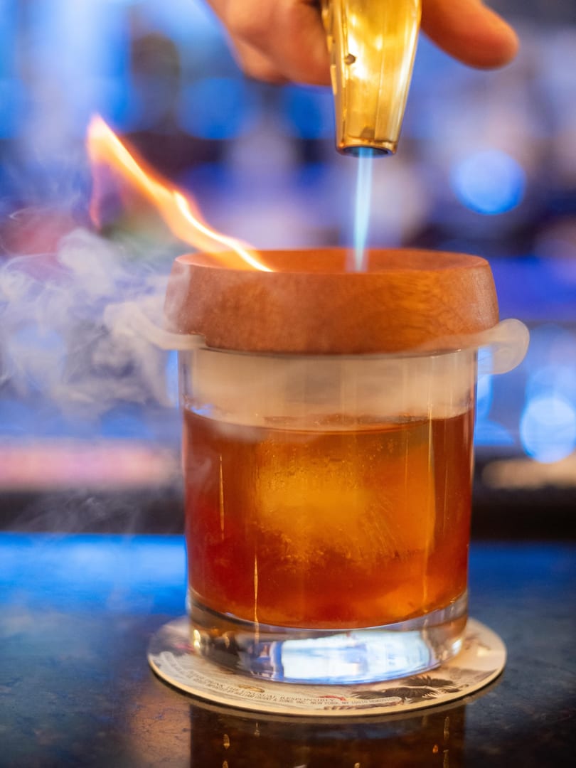 A bartender uses a torch to smoke a cocktail in a glass with a large ice cube.