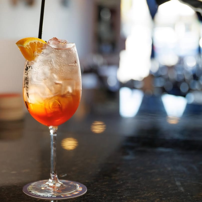 A vibrant orange cocktail with ice and an orange slice sits on a dark bar.