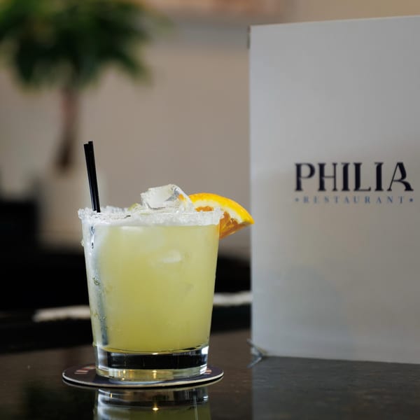 A Philia Restaurant menu stands behind a margarita with a salted rim, orange slice, and black straws.