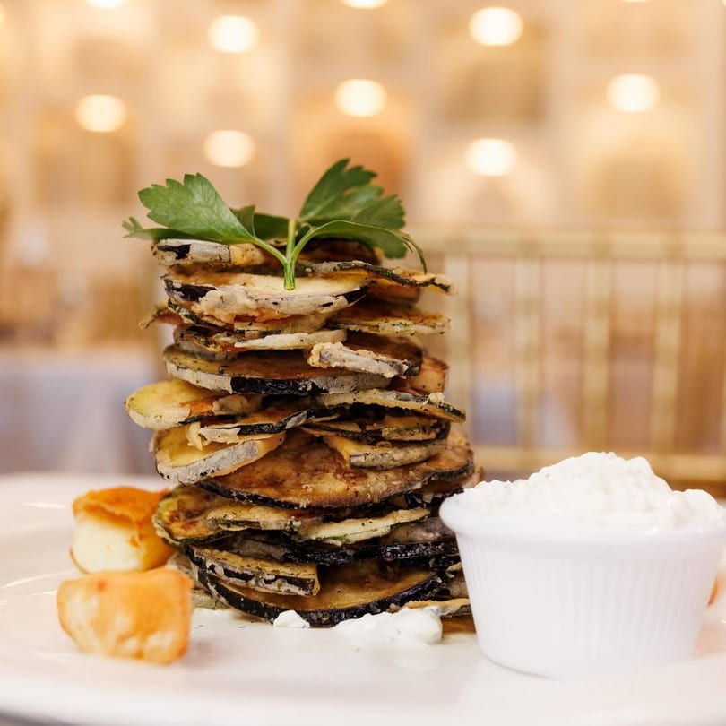 Tall stack of fried zucchini and eggplant slices with a side of white dip and cheese.