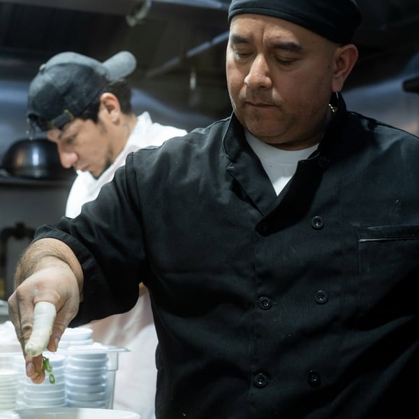 A chef in a black uniform garnishes plates of food with green onions in a busy kitchen.