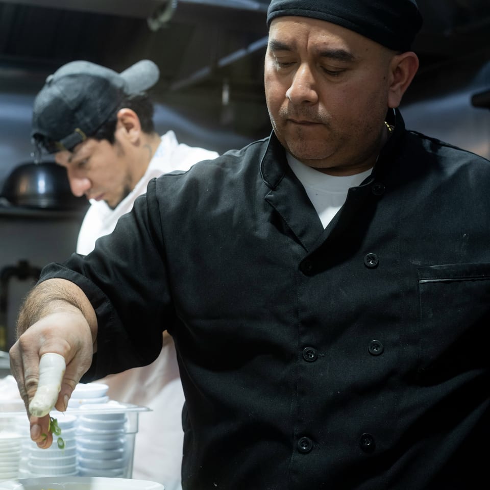 A chef in a black uniform garnishes plates of food with green onions in a busy kitchen.