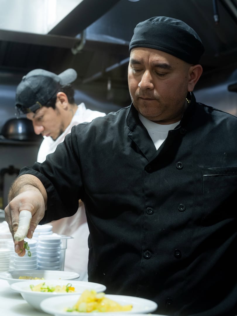 A chef in a black uniform garnishes plates of food with green onions in a busy kitchen.