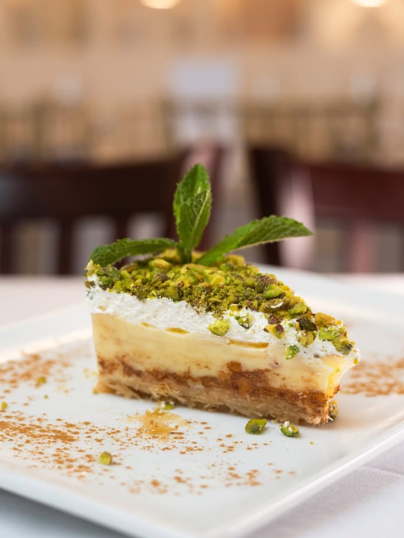 Slice of dessert with layers of pastry, cream, and pistachios, garnished with mint and cinnamon.